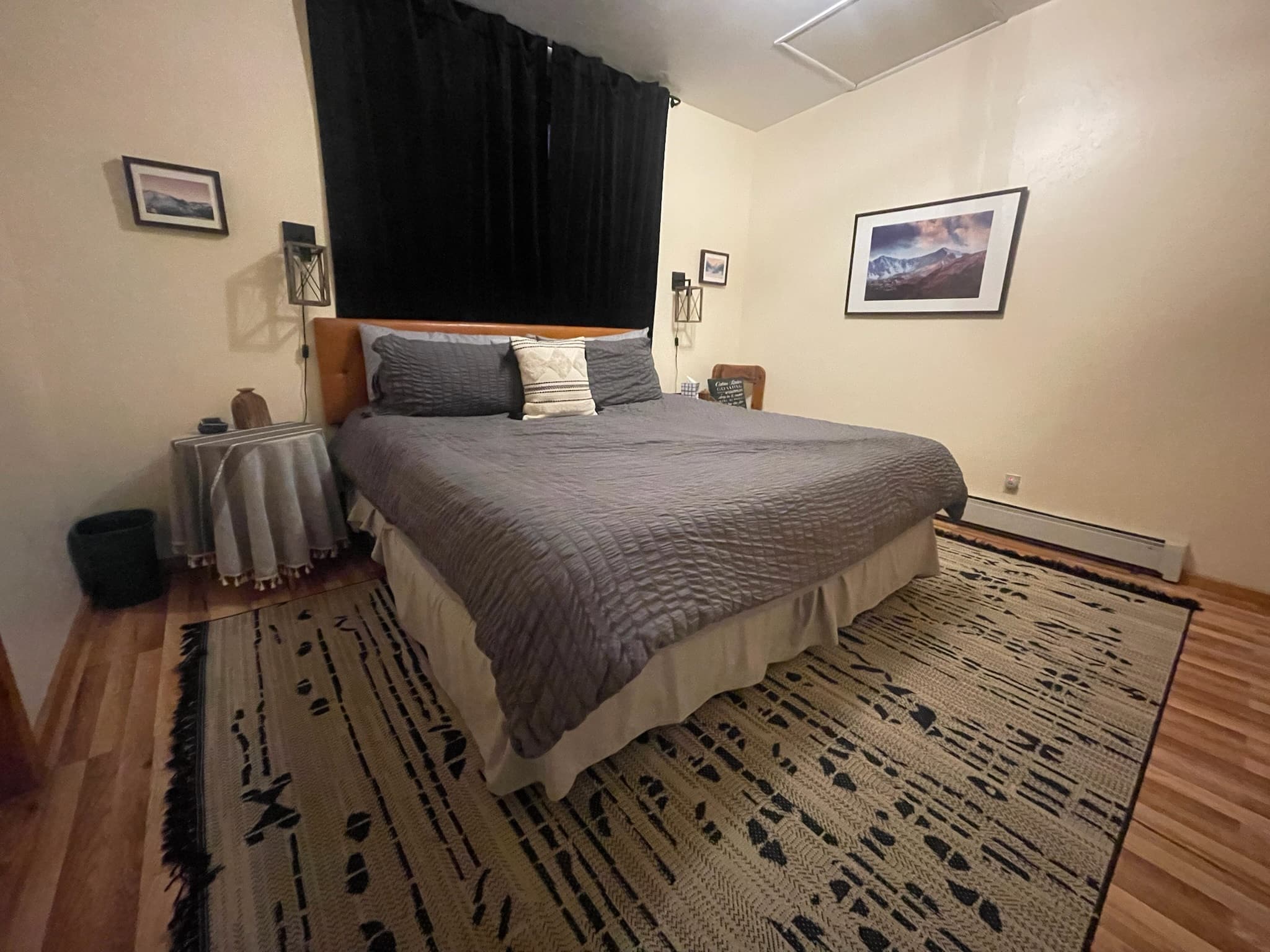 Cozy bedroom with a grey bed, black curtains, mountain landscape art, and a patterned rug.