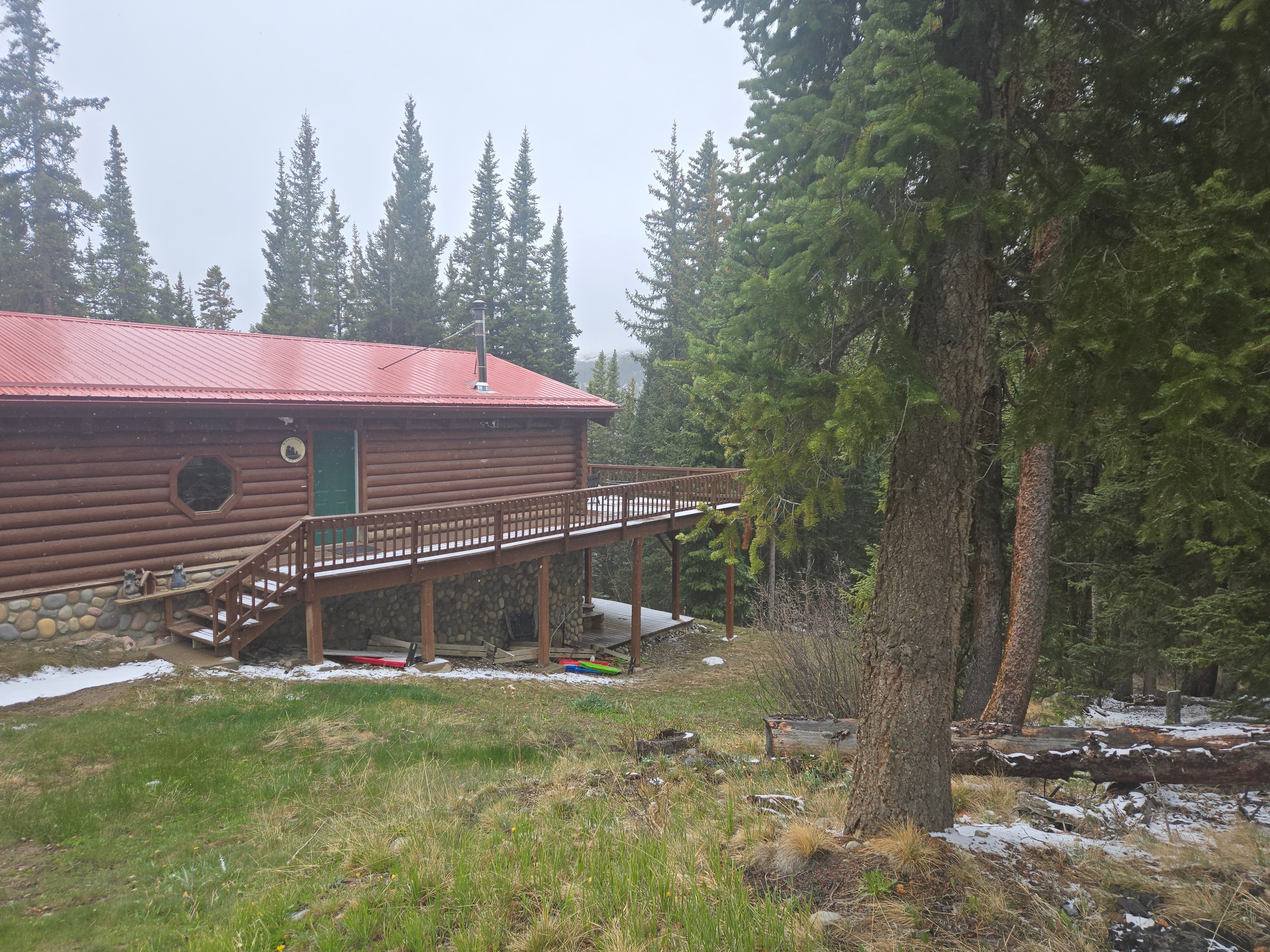 Log cabin with a red metal roof and wooden deck surrounded by snowy evergreen trees.