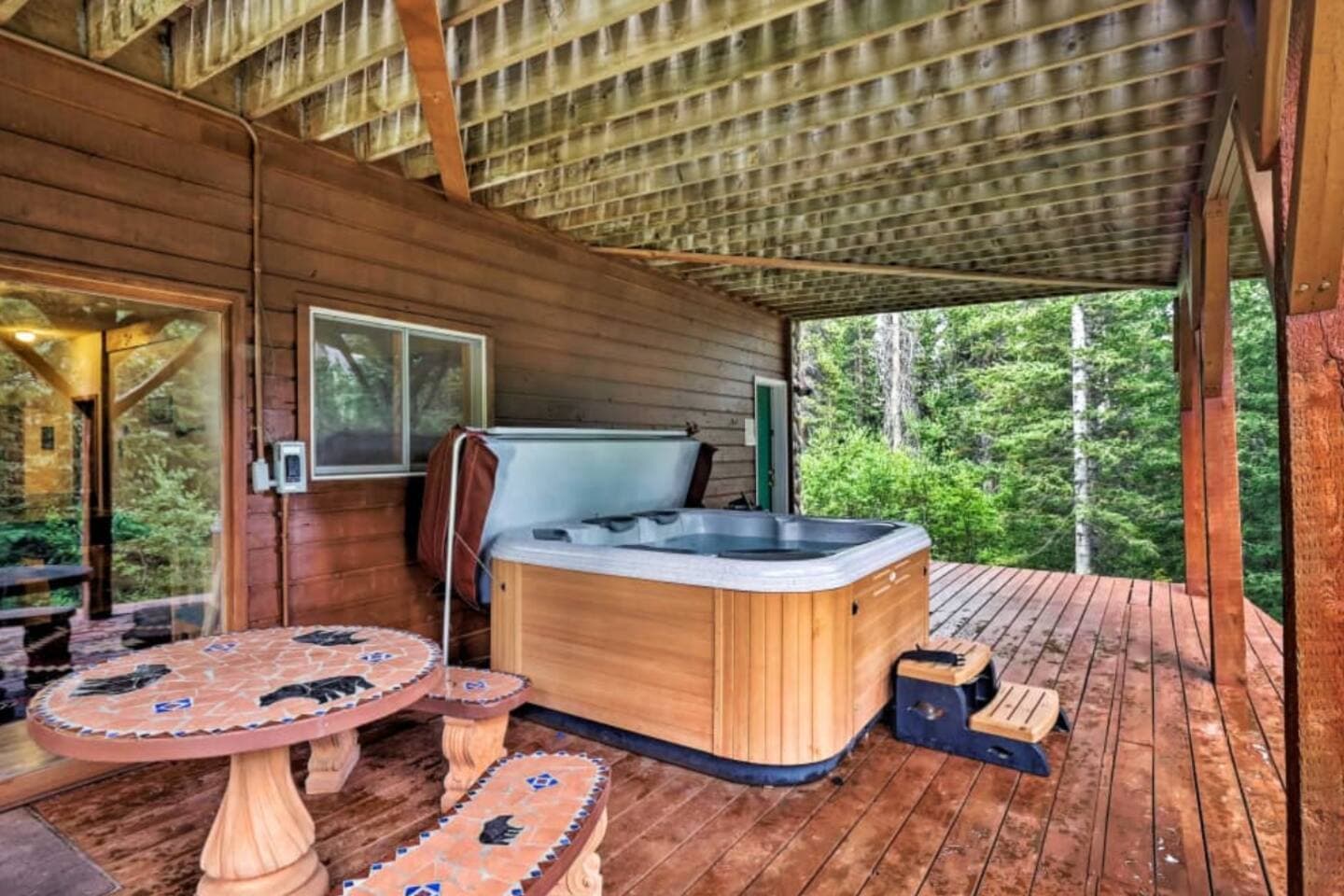 Hot tub and bear-themed table on a covered wooden deck overlooking a lush forest.