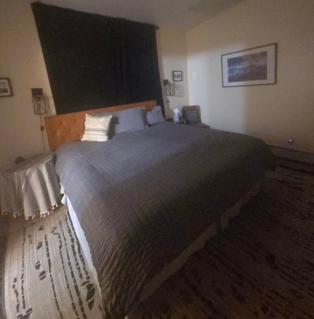 Large bed with grey textured comforter, leather headboard, and black fabric backdrop in a bedroom.