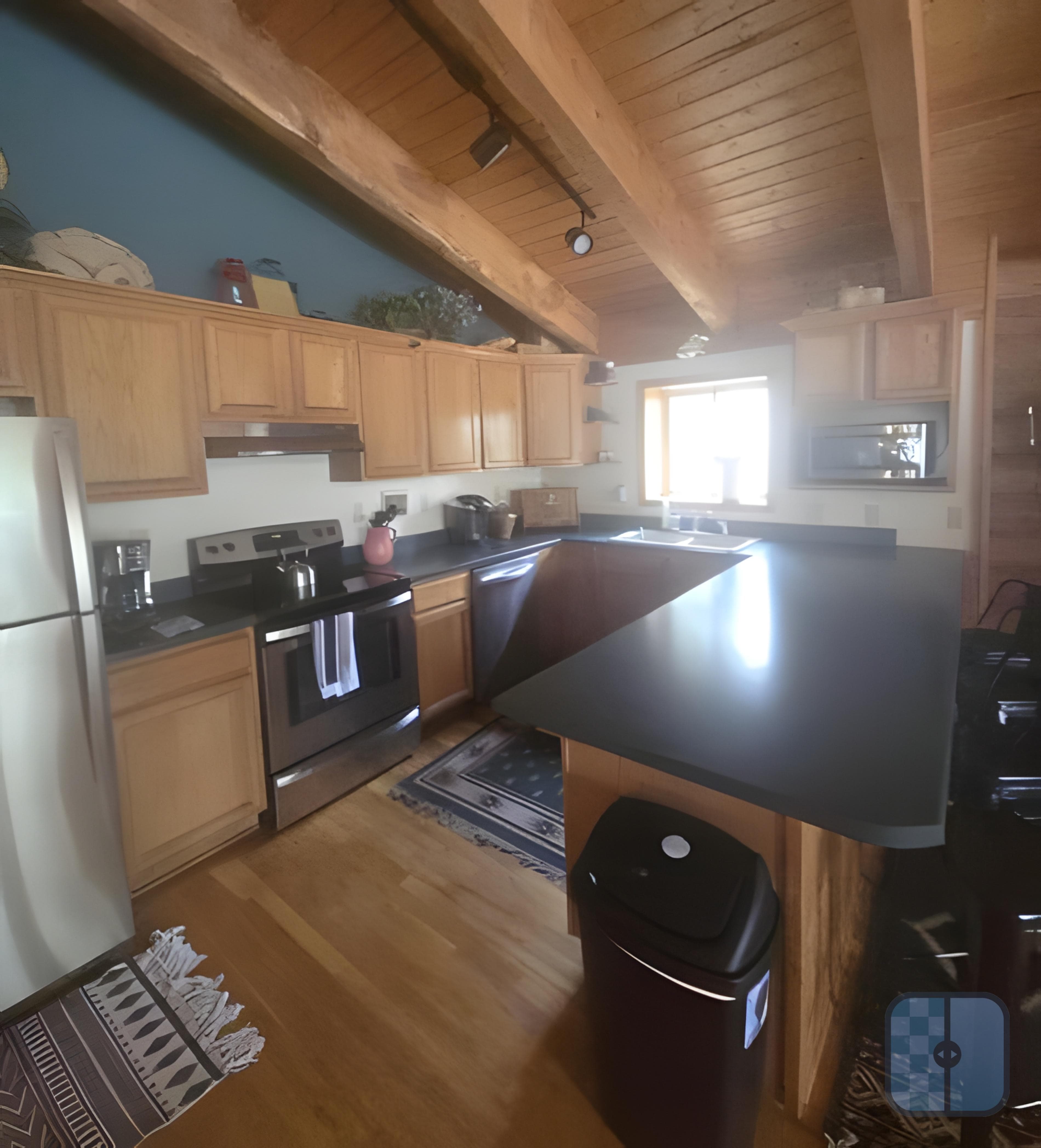 Rustic kitchen featuring light wood cabinets, dark countertops, stainless steel appliances, and exposed wooden beams.