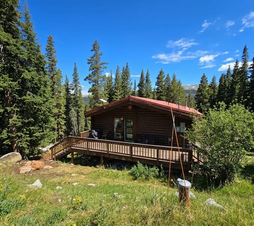 Rustic log cabin with a red roof and wooden deck surrounded by tall evergreen trees.