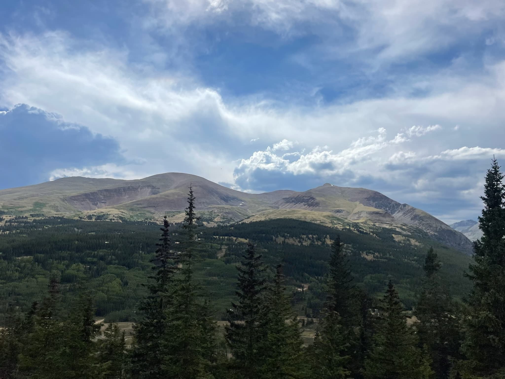 Rolling mountain peaks rise above a dense evergreen forest under a dramatic, cloud-filled sky.