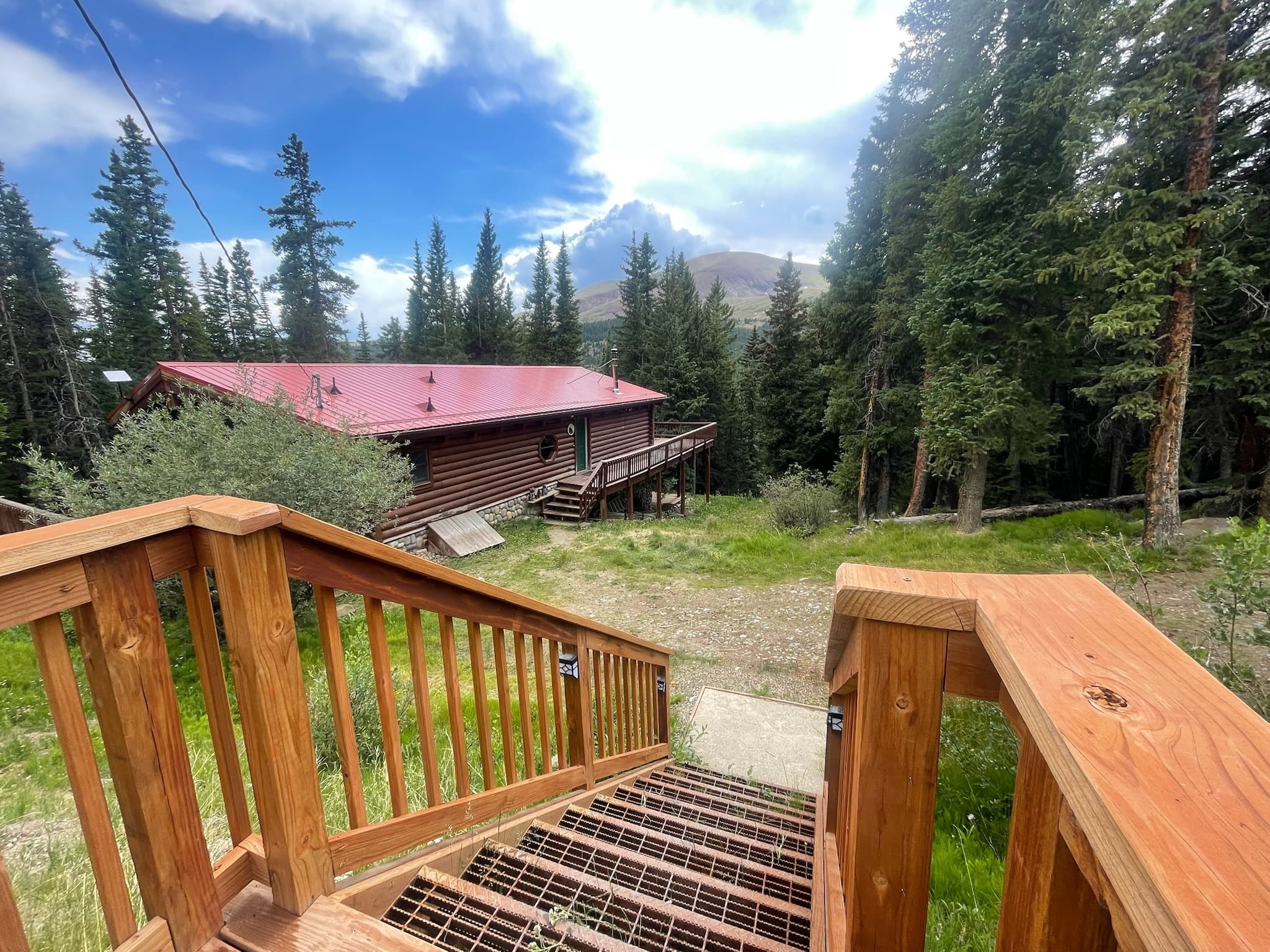 Log cabin with a red roof nestled in a pine forest, viewed from wooden stairs.
