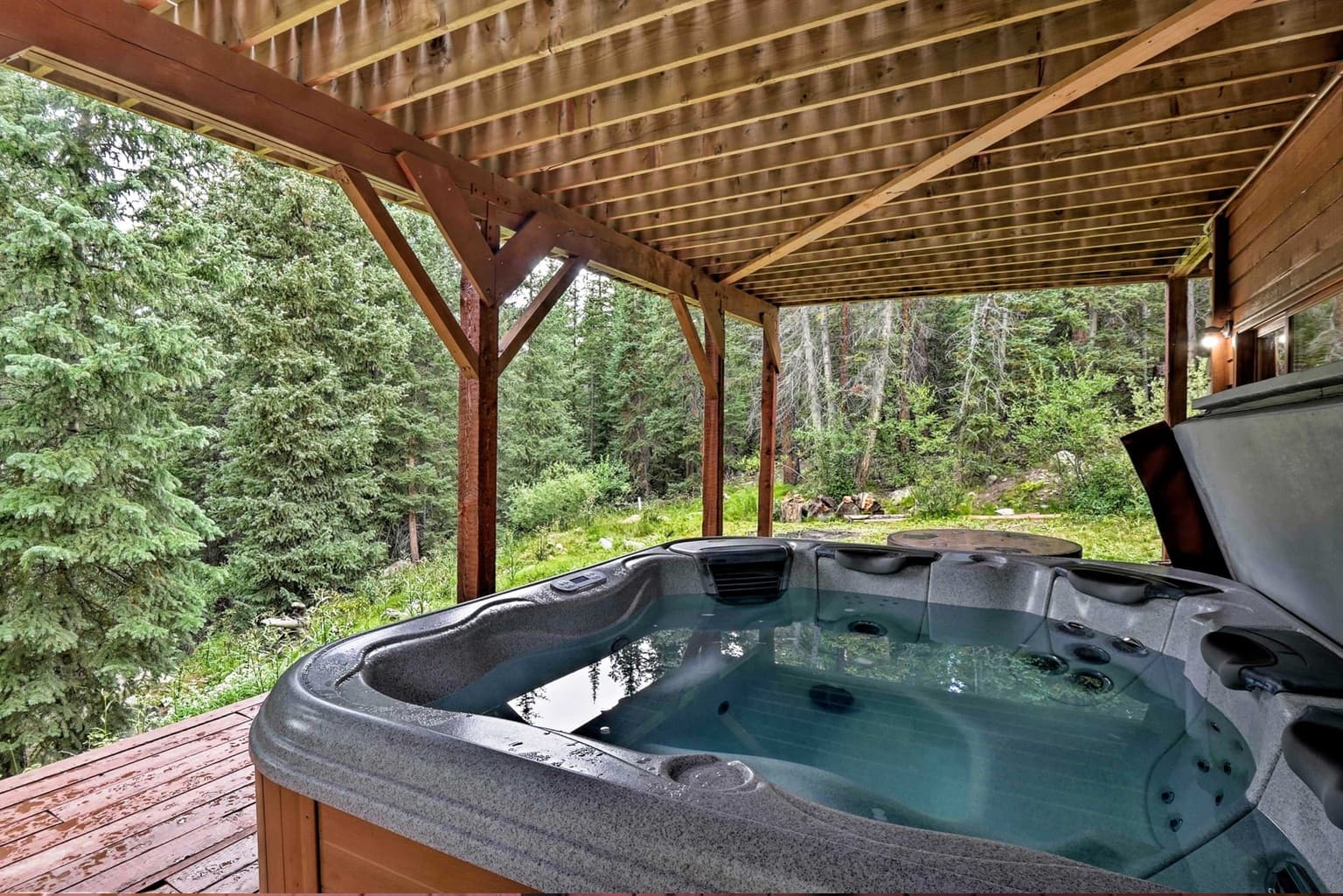 Hot tub on a wooden deck under a pergola overlooking a lush evergreen forest.