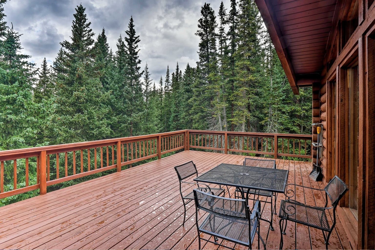Wooden deck with black metal furniture overlooking a dense evergreen forest under overcast skies.