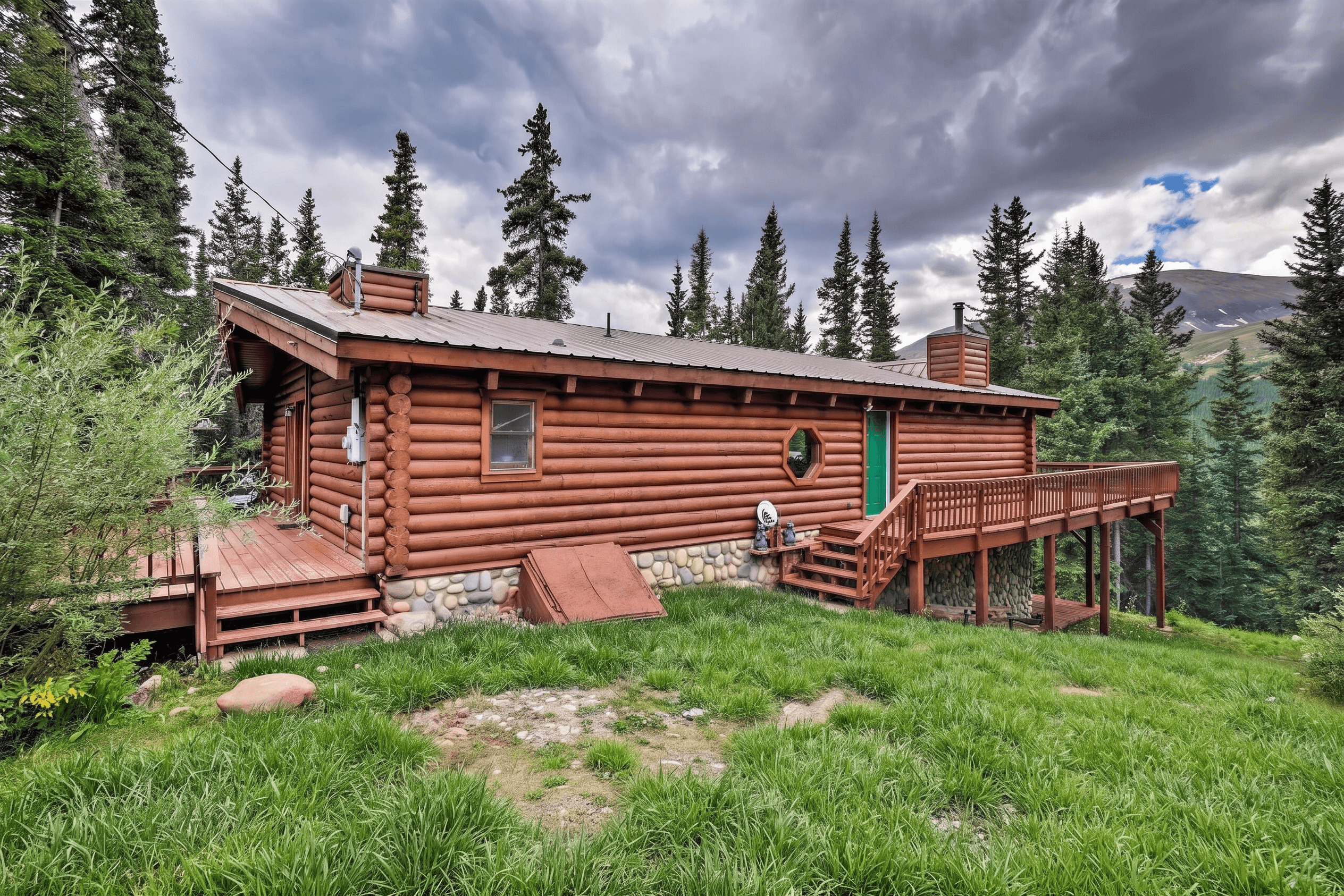 Rustic log cabin with a wooden deck surrounded by tall pine trees under cloudy skies.